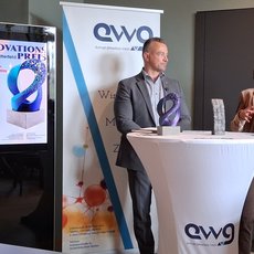 Drei Personen auf einer Bühne: Ein Mann mit verschränkten Armen, ein Mann in Anzug, der hinter einem Podium mit Mikrofon spricht, und eine Frau in Businesskleidung, die ebenfalls spricht. Im Hintergrund ein Banner mit „Innovationspreis“ und dem Logo „ewg“.