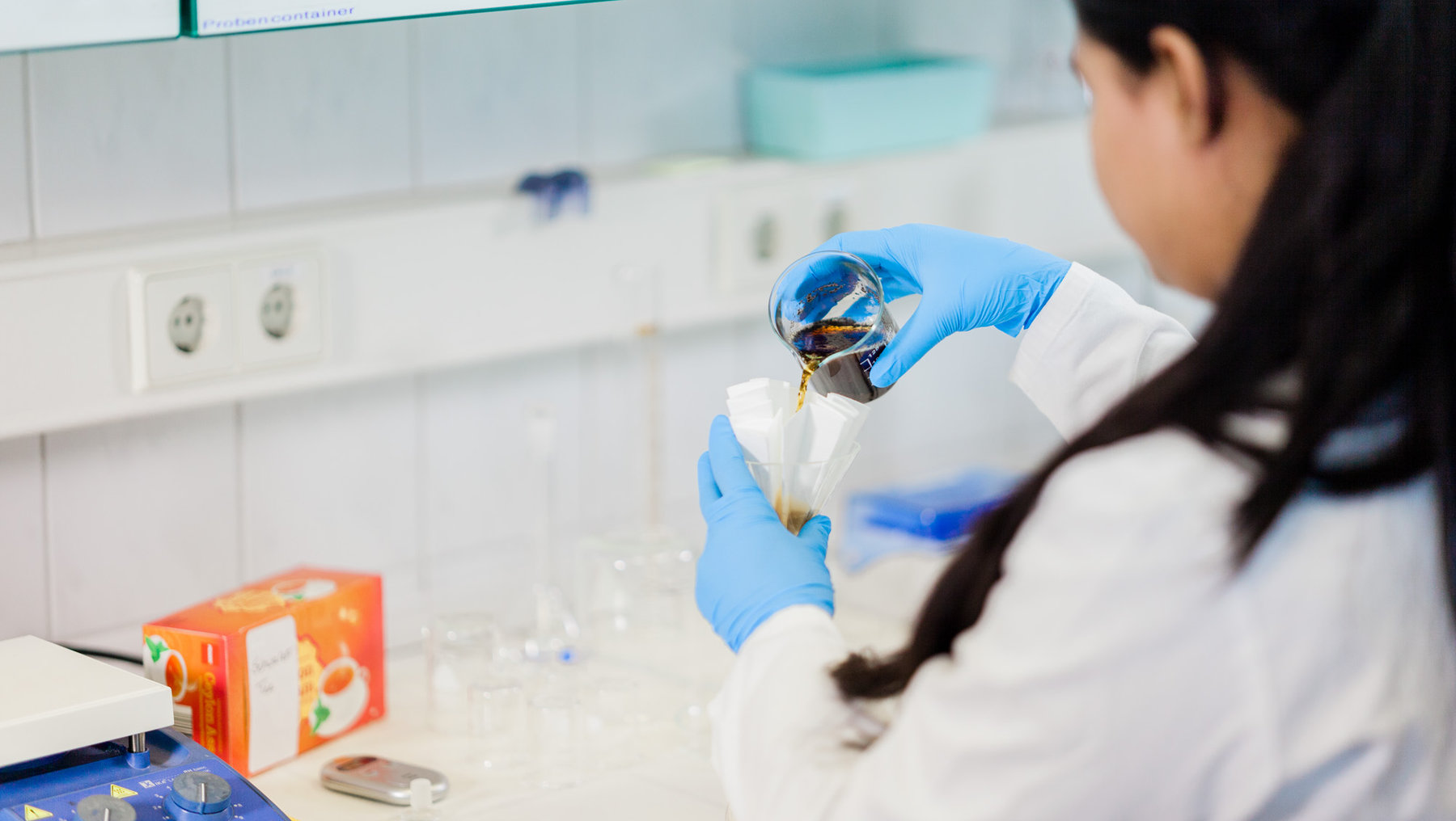 Eine Person in weißem Laborkittel und blauen Handschuhen überführt eine dunkle Flüssigkeit aus einem Glasbehälter in ein Reagenzglas, das in einem Halter steckt. Im Hintergrund sind Laborgeräte und Chemikalienbehälter zu sehen.