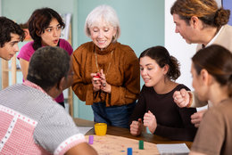 Eine ältere Frau erklärt lächelnd einer Gruppe junger Erwachsener an einem Tisch spielerisch etwas, während alle konzentriert mit den Händen auf ein Brettspiel oder eine Aktivität zeigen.