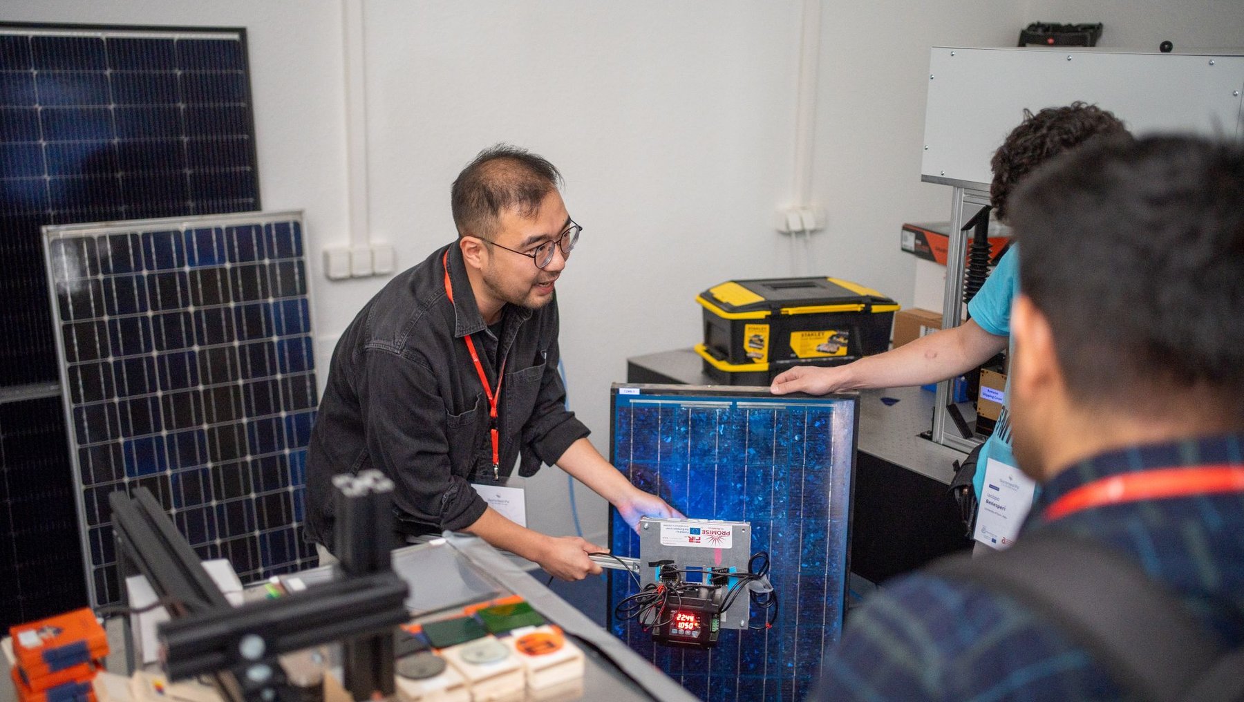 Zwei Personen arbeiten in einem Labor mit Solarmodulen und technischer Ausrüstung. Ein Mann erklärt oder zeigt auf ein kleines Gerät, das mit einem blauen Kasten verbunden ist, während der andere aufmerksam zuhört. Im Hintergrund sind weitere Solarpaneele und Werkzeuge zu sehen.