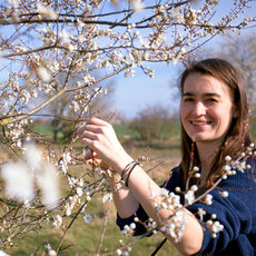 A woman with long hair smiles while touching the blossoms of a flowering tree in an open field. The tree is in full bloom with white flowers, and the background shows a green landscape under a clear sky.