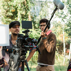 A film crew is working outdoors. One person operates a camera on a tripod, another holds a boom microphone, and a third person adjusts a light reflector. They appear to be filming a scene in a natural setting with trees in the background.