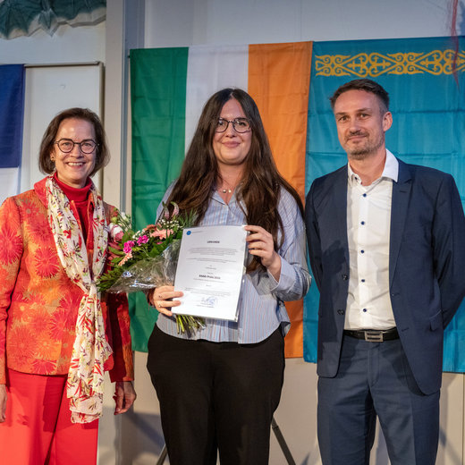 Drei Personen stehen lächelnd auf einer Bühne. Eine Frau in der Mitte hält einen Blumenstrauß und eine Urkunde mit der Aufschrift DAAD-Preis 2026. Links und rechts stehen zwei weitere Personen, eine mit Schal und eine im Anzug. Im Hintergrund hängen Flaggen und ein Banner.