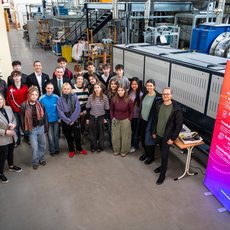 Gruppe von etwa 20 Personen steht lächelnd vor einem großen technischen Gerät, vermutlich einer Maschine oder Anlage, in einem Industrieumfeld. Dahinter sind Werkzeuge und Maschinen zu sehen. Rechts steht ein Banner mit dem Schriftzug „Festo Engineering“.
