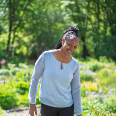 Eine lächelnde Person mit Zöpfen steht in einem sonnigen Garten, berührt zart eine blühende Pflanze mit blauen und violetten Blüten, umgeben von üppiger grüner Vegetation.