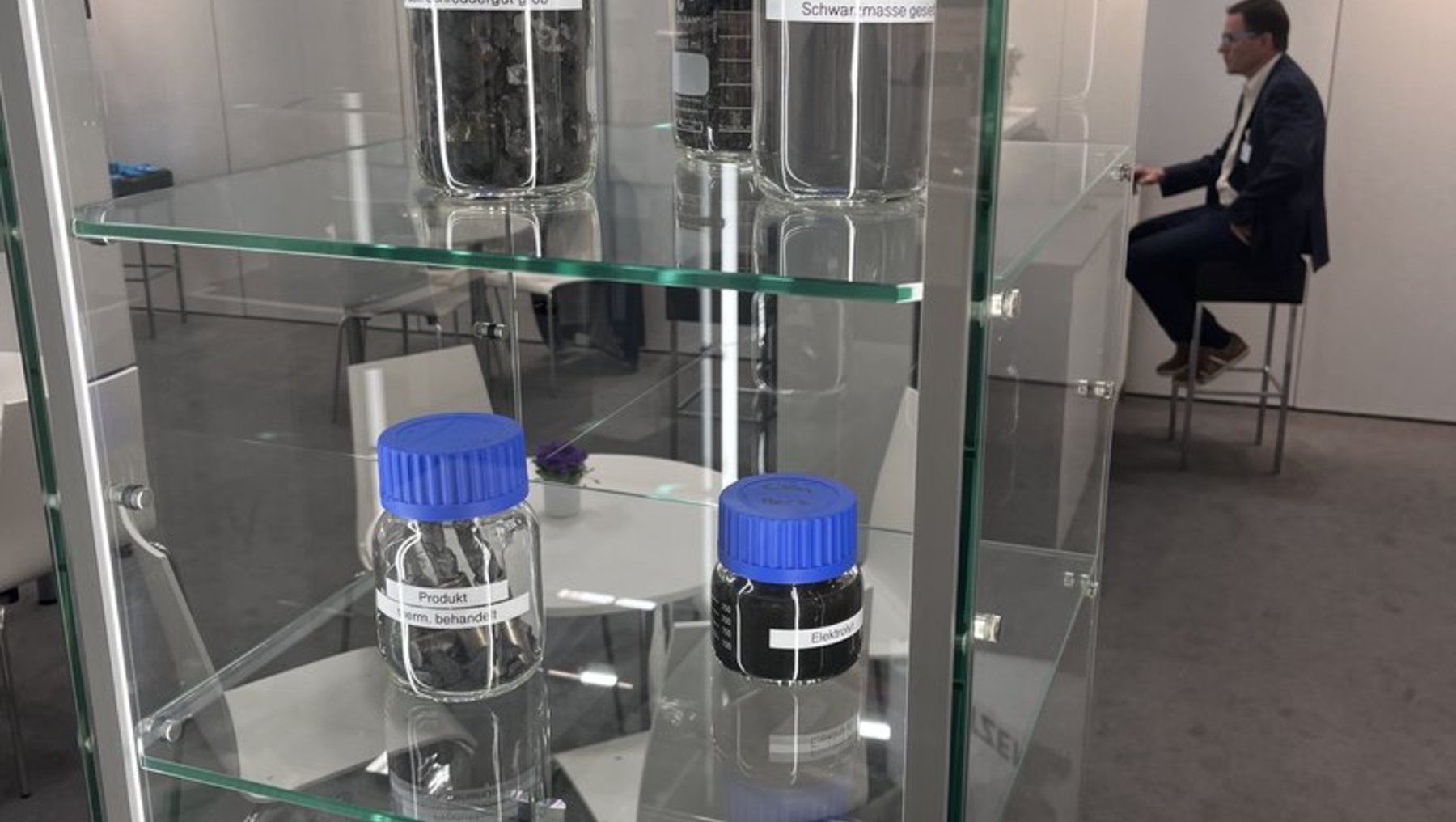 Glassvitrine mit vier durchsichtigen Behältern, gefüllt mit dunklem, körnigem Material, vermutlich Kaffee- oder Teebohnen, jeweils mit blauen Deckeln. Darunter liegt ein Tablet mit einer farbigen Anzeige und einem Stift. Im Hintergrund sitzt eine Person in Anzug.