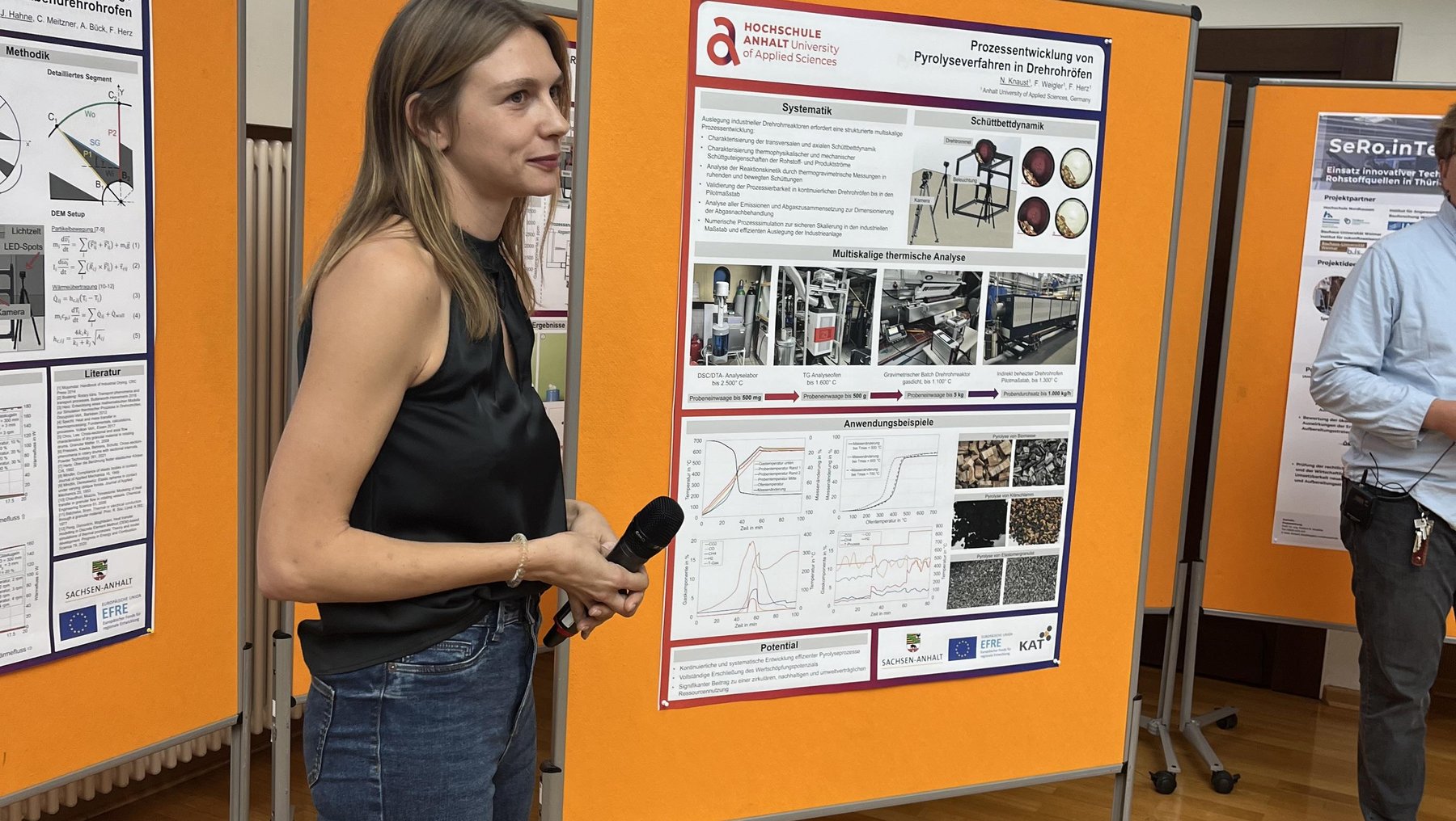 Eine Person steht vor einem orangefarbenen Poster mit wissenschaftlichem Inhalt, hält ein Mikrofon und präsentiert. Das Poster zeigt Grafiken, Fotos und Text in Russisch und Englisch zu einem technischen oder archäologischen Thema. Im Hintergrund weitere Poster und eine Person im Hintergrund.