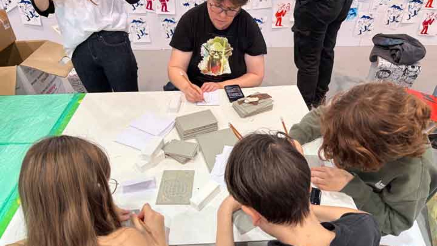 Eine Gruppe von vier Personen sitzt an einem Tisch und arbeitet gemeinsam an einem kreativen Projekt mit Materialien wie Pappe, Stiften und kleinen Modellen. Eine Person im Mittelpunkt erklärt oder zeigt etwas, während die anderen konzentriert mitmachen.
