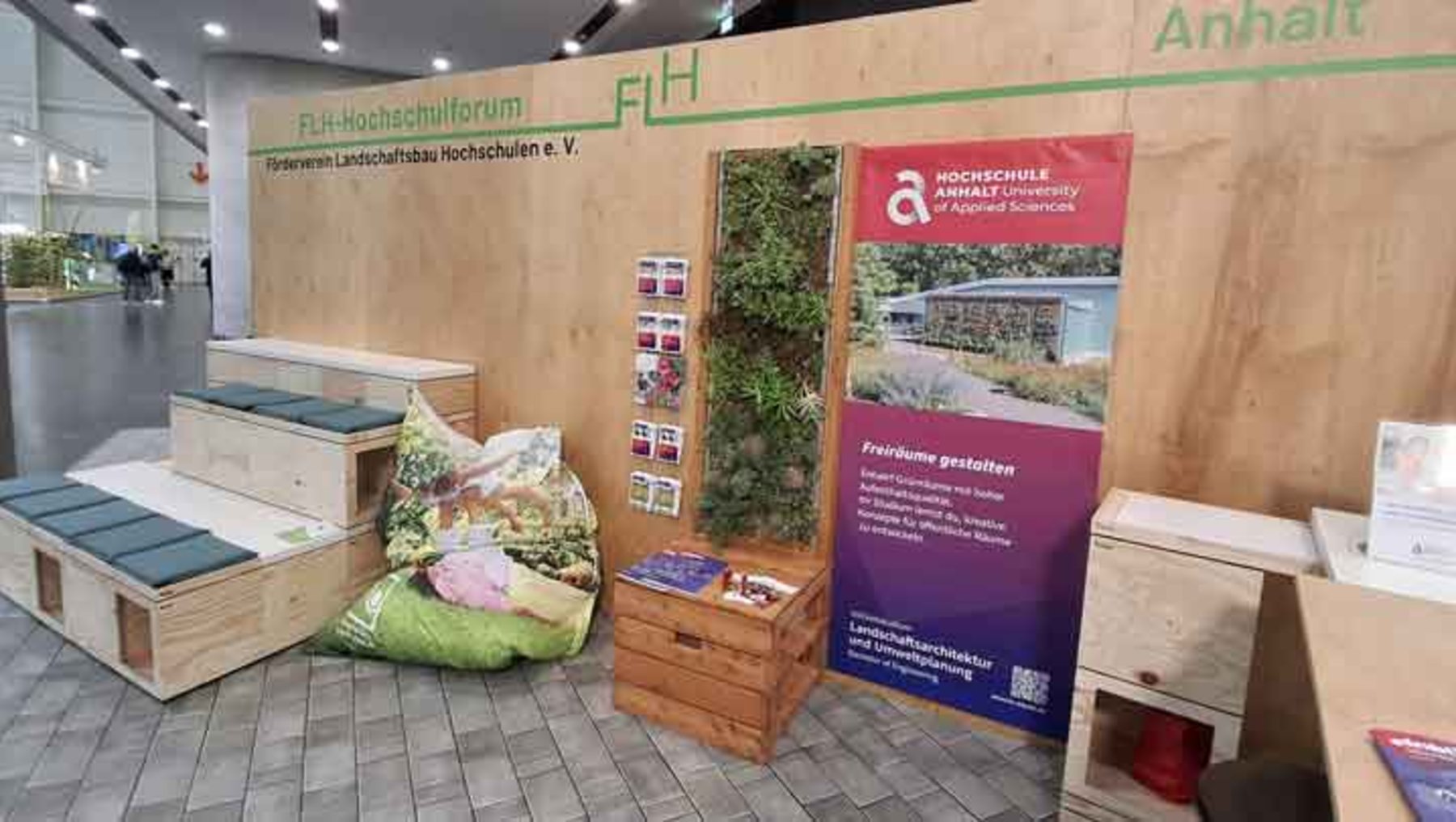 Ausstellungstand der FH Hochschule Rheinland-Pfalz mit Infomaterialien zu nachhaltigem Bauen und Landschaftsarchitektur. Gezeigt werden Modelle von Hochbeeten und Möbeln aus Holz, umgeben von Pflanzen. Plakate und Broschüren liegen bereit.