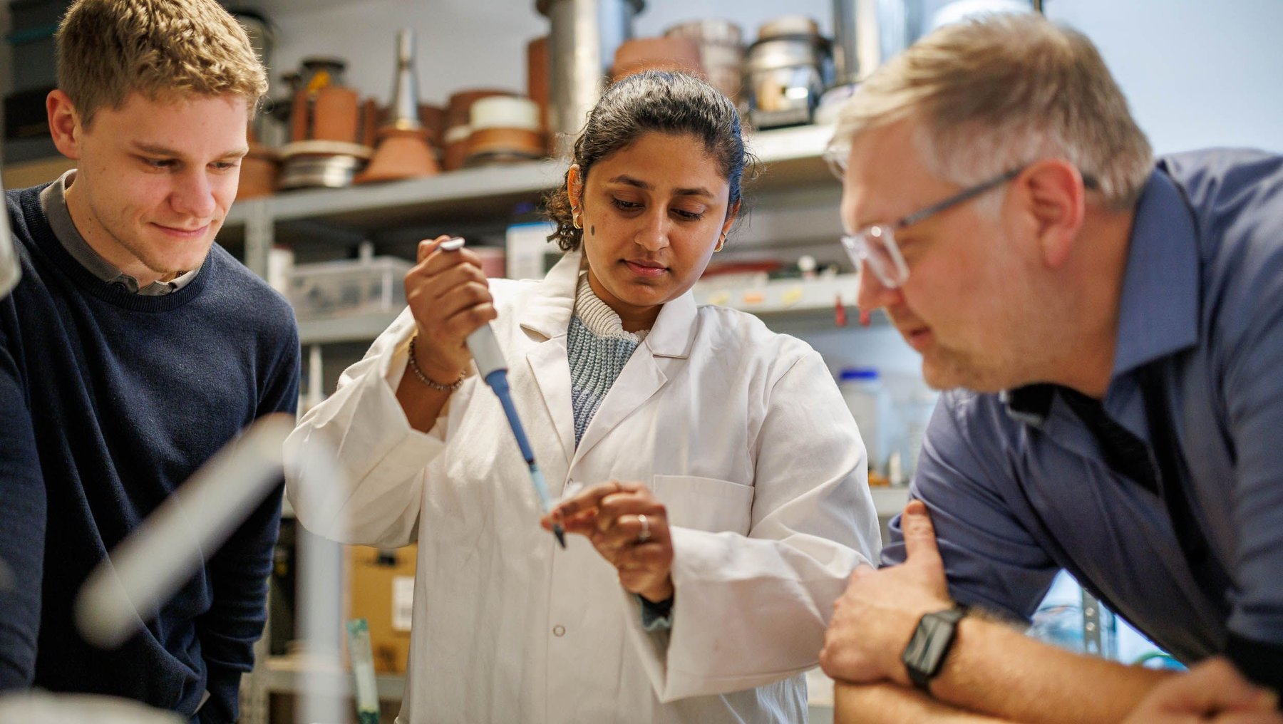 Drei Personen arbeiten in einem Labor. Eine Frau in einem weißen Laborkittel hält eine Pipette und wird von zwei Männern beobachtet, die sich auf die Arbeit konzentrieren. Im Hintergrund sind Laborgeräte und Chemikalien zu sehen.