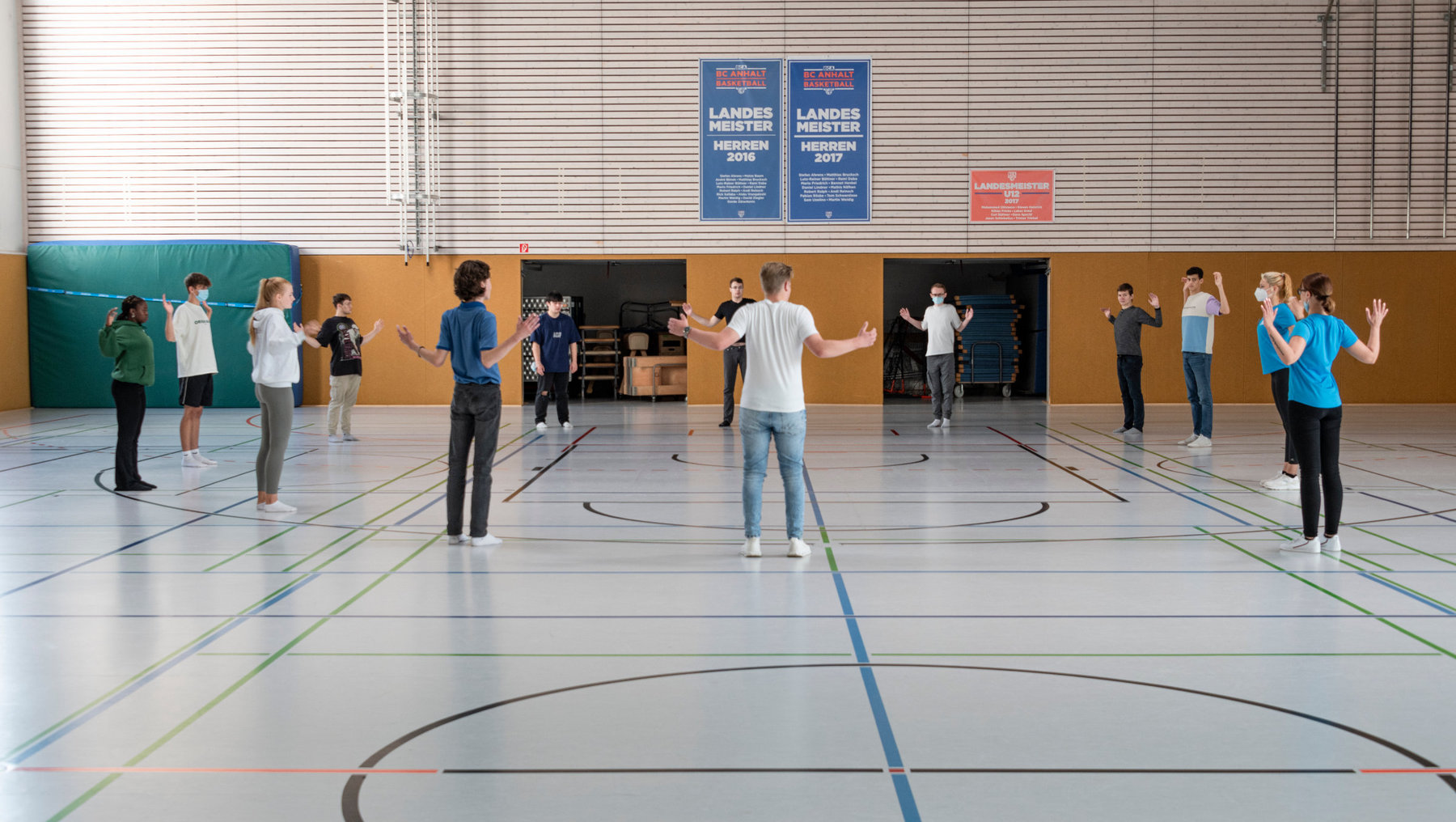 Gruppe von Personen in einer Sporthalle, die in einem Kreis stehen und gemeinsam eine Übung mit Armbewegungen durchführen, vermutlich zur Koordination oder zum Warm-up.