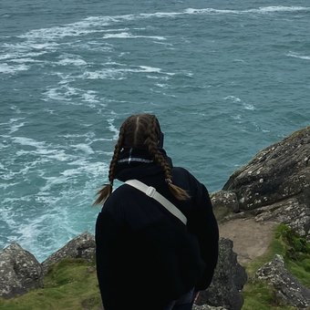 Eine Person steht an einer steilen Klippe und blickt auf das weite, leicht bewegte Meer. Im Vordergrund ragen große Felsen aus dem Gras hervor, während der Himmel grau und neblig wirkt.