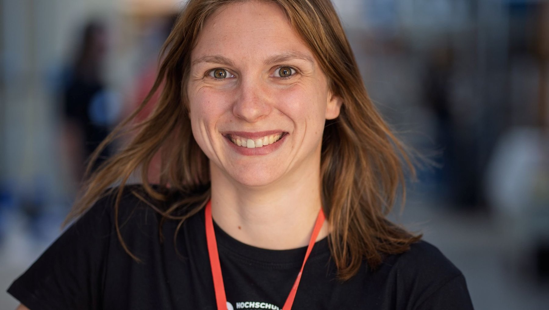 Lächelnde Frau mit mittellangem, braunem Haar, die ein schwarzes T-Shirt trägt und ein rotes Band mit Logo um den Hals hat. Sie steht vor einem unscharfen Hintergrund mit Menschen im Industrielabor der Hochschule Anhalt und hält sich mit einer Hand am Arm fest.