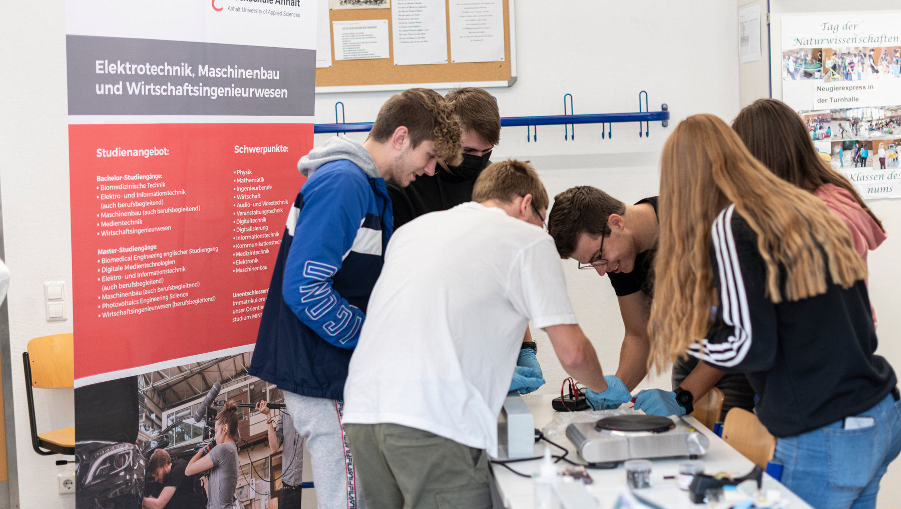 Eine Gruppe von fünf Personen, darunter vier Männer und eine Frau, bespricht gemeinsam ein technisches Projekt an einem Tisch mit elektronischen Bauteilen. Im Hintergrund steht ein Infoposter zu Studiengängen in Elektrotechnik, Maschinenbau und Wirtschaftsingenieurwesen.