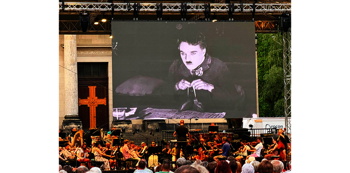 Großes Open-Air-Konzert mit Orchester und Chor vor einer Bühne, auf der ein Schwarz-Weiß-Film von Charlie Chaplin mit ihm in einer traurigen Pose beim Geigenspiel projiziert wird.