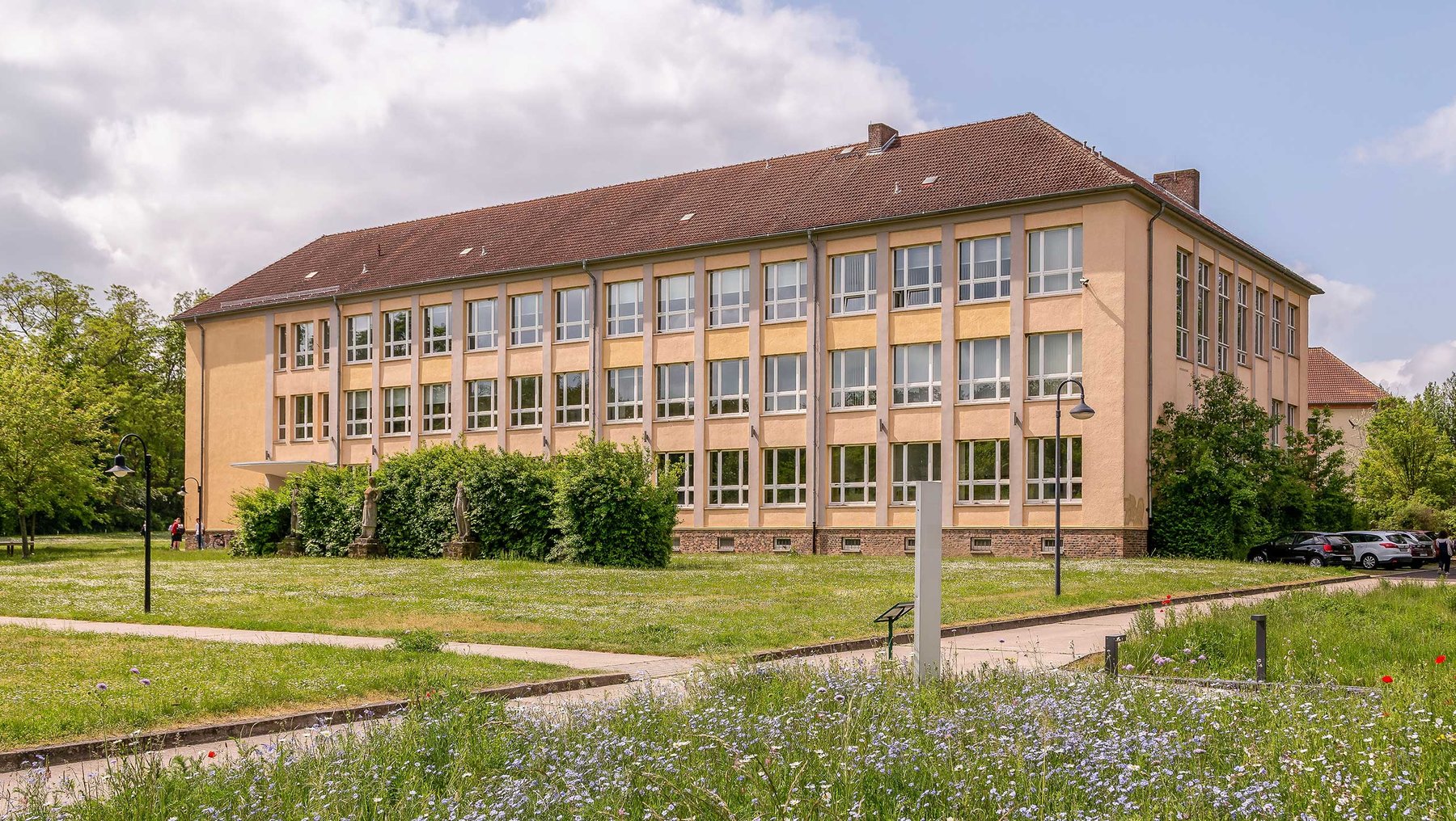 Ein großes, zweistöckiges Schulgebäude mit vielen Fenstern, umgeben von einem grünen Rasen und Büschen. Der Himmel ist teilweise bewölkt. Im Vordergrund sind einige Blumen zu sehen.
