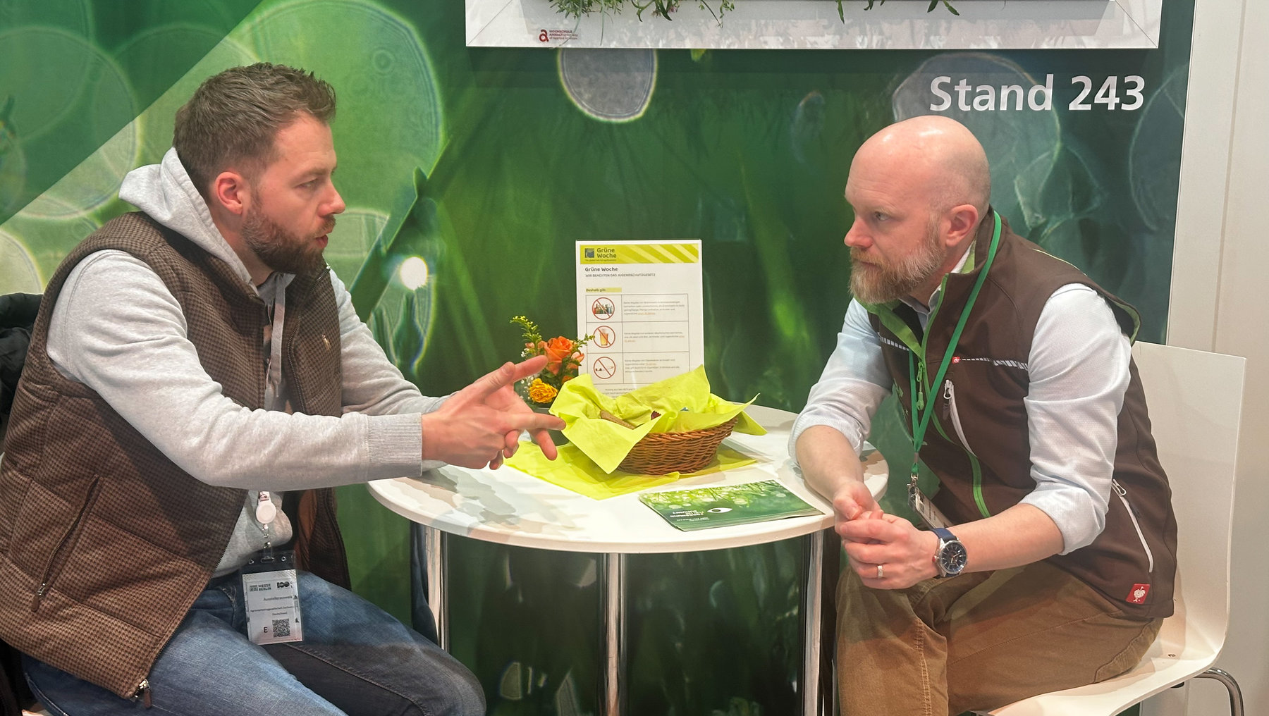 Zwei Männer sitzen an einem Messestand mit der Nummer 243 und besprechen sich über eine kleine Pflanze in einem Korb mit gelber Verpackung, umgeben von Informationsmaterial zu „Grüner Woche“.