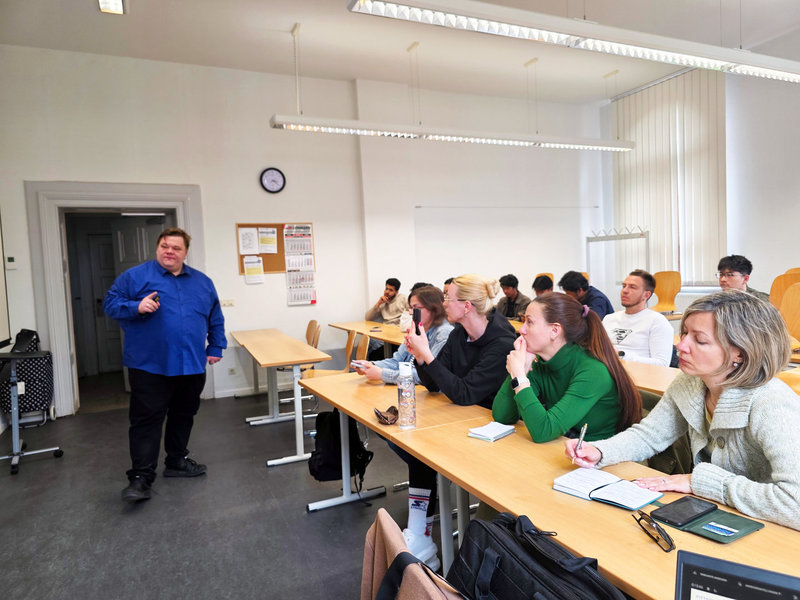 Ein Mann steht in einem hellen Seminarraum und hält eine Präsentation vor einer Gruppe von Zuhörern, die an Tischen sitzen und Notizen machen. Der Raum wirkt wie ein klassischer Unterrichts- oder Schulungsbereich mit Tischen, Stühlen und einer Tür im Hintergrund.