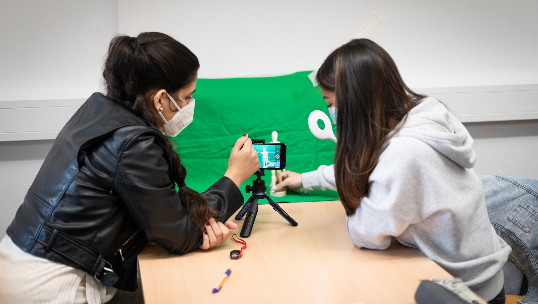 Zwei Personen sitzen an einem Tisch und filmen sich gegenseitig mit einem Smartphone auf einem Stativ vor einem grünen Hintergrund mit weißem Logo. Beide tragen Masken.