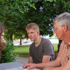 Three people are sitting outdoors at a table, engaged in a discussion. They are looking at a document together. The setting appears to be a park with trees and buildings in the background. The individuals seem to be having a serious conversation.