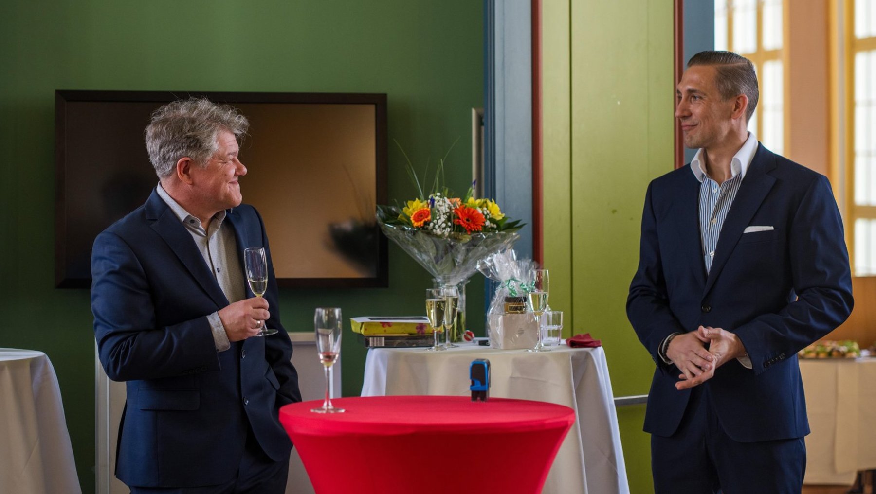 Zwei Herren in Anzügen stehen in einem Raum mit gedecktem Tisch. Der Mann links hält ein Glas Wein und spricht, während der Mann rechts zuhört. Auf dem Tisch stehen eine Blumenkomposition und Gläser.