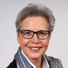 A woman with short gray hair, wearing glasses, a patterned scarf, and a dark top, smiling at the camera. The background is plain and light-colored.