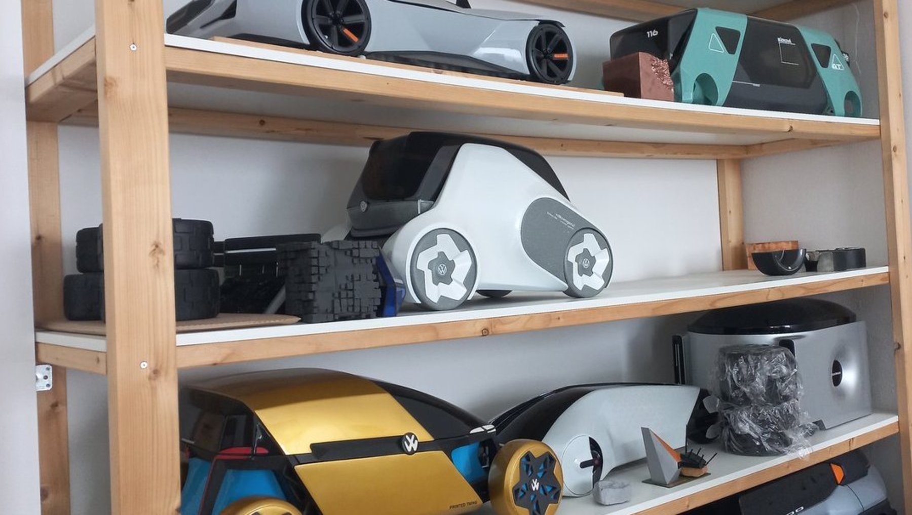 Drei Regalböden mit futuristischen Modellautos in verschiedenen Farben und Designs: oben silbernes Auto mit schwarzer Front, mittig weißes Auto mit blauen und schwarzen Akzenten, unten gelbes Auto mit blauen Rädern und silbernes Auto mit grauer Abdeckung.