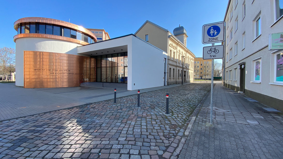 Moderne Gebäude mit weißer Fassade und holzverkleideter Rundung, die ein offenes Eingangsportal zeigt. Vor dem Gebäude eine gepflasterte Straße mit Fahrradzonen-Schild und Parkplatzmarkierungen. Im Hintergrund historische Backsteingebäude.