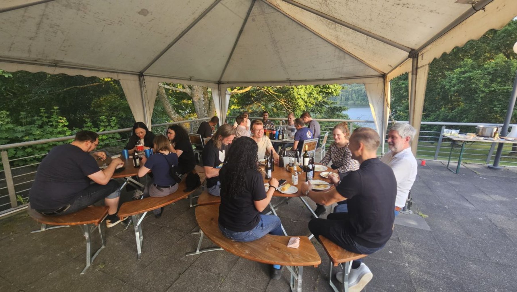 Gruppe von Menschen sitzt an runden Holztischen unter einem Zeltdach, isst und unterhält sich in entspannter Atmosphäre. Im Hintergrund ist ein Geländer und grüne Natur zu sehen.