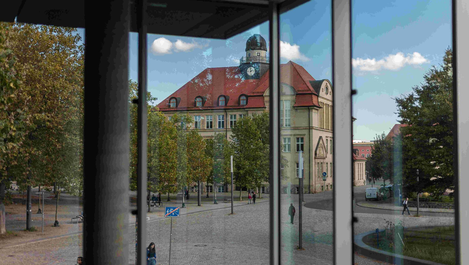 Ein Blick durch ein Fenster auf einen Platz mit einem historischen Gebäude, das eine Uhr und ein rotes Dach hat. Im Vordergrund ist eine Straße mit Fußgängern und Bäumen zu sehen. Im Hintergrund befindet sich ein Brunnen. Der Himmel ist blau mit einigen Wolken.