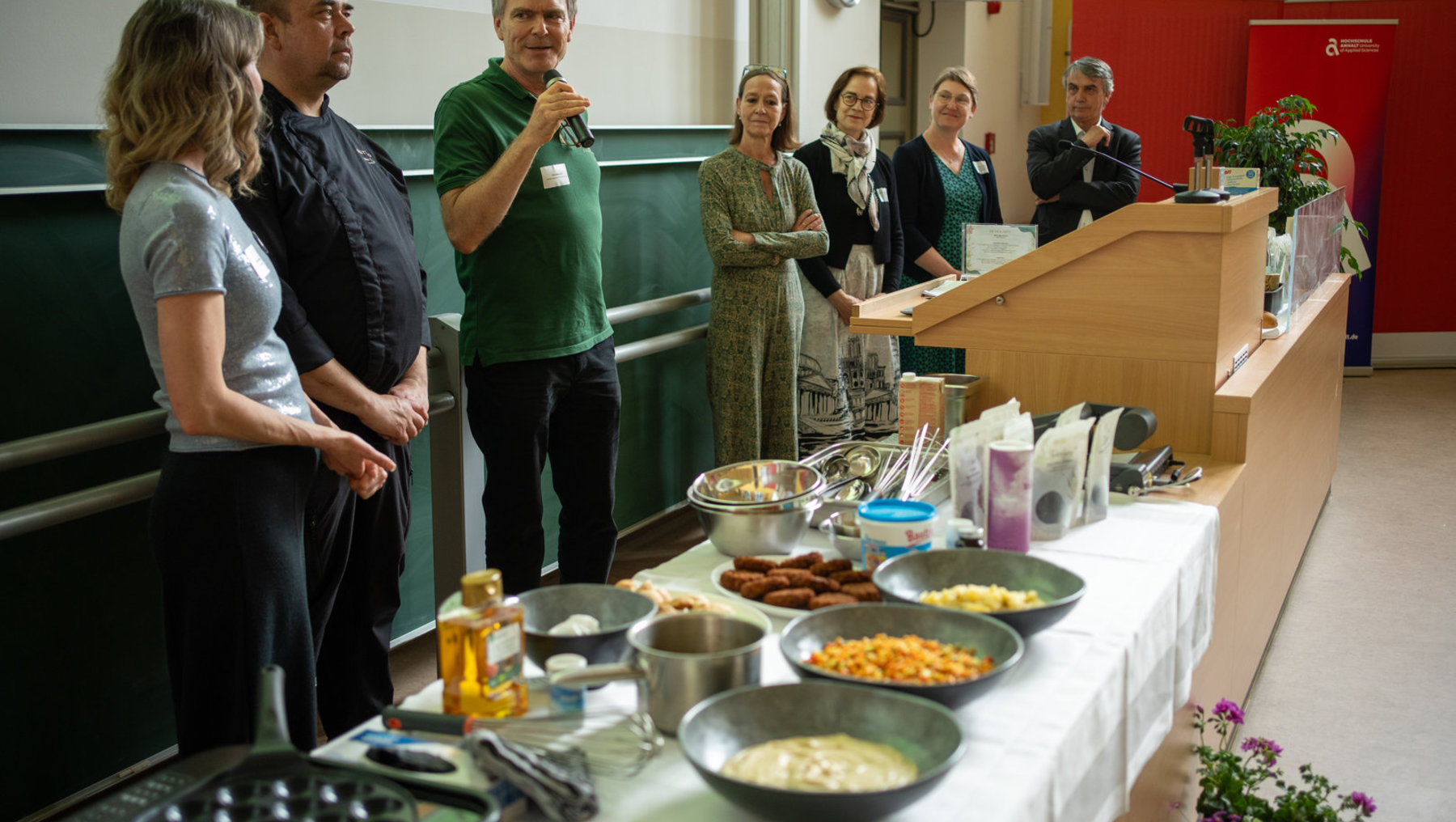 Ein Mann spricht mit Mikrofon an einem Rednerpult, während mehrere Personen im Hintergrund zuhören. Vor ihm steht ein Buffet mit verschiedenen Snacks wie Käse, Obst, Nüsse und Süßigkeiten sowie Küchenutensilien.