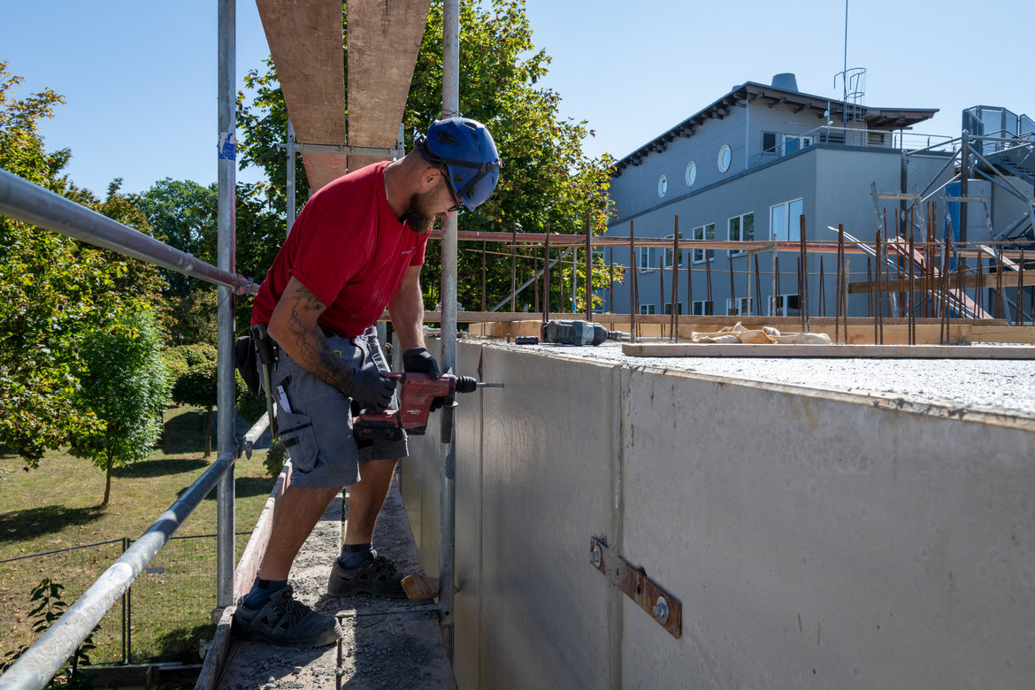 Ein Bauarbeiter mit Schutzhelm und Sicherheitsausrüstung arbeitet auf einer Baustelle an einer Betonwand. Er hält einen Bohrer und befestigt oder bearbeitet etwas an der Wand. Im Hintergrund ist ein Gebäude im Rohbau zu sehen.