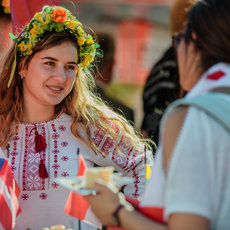 Eine lächelnde Frau mit einem Blumenkranz aus gelben, grünen und roten Blüten auf dem Kopf, die traditionelle Stickmuster auf ihrem weißen Oberteil trägt und kleine Flaggen hält.
