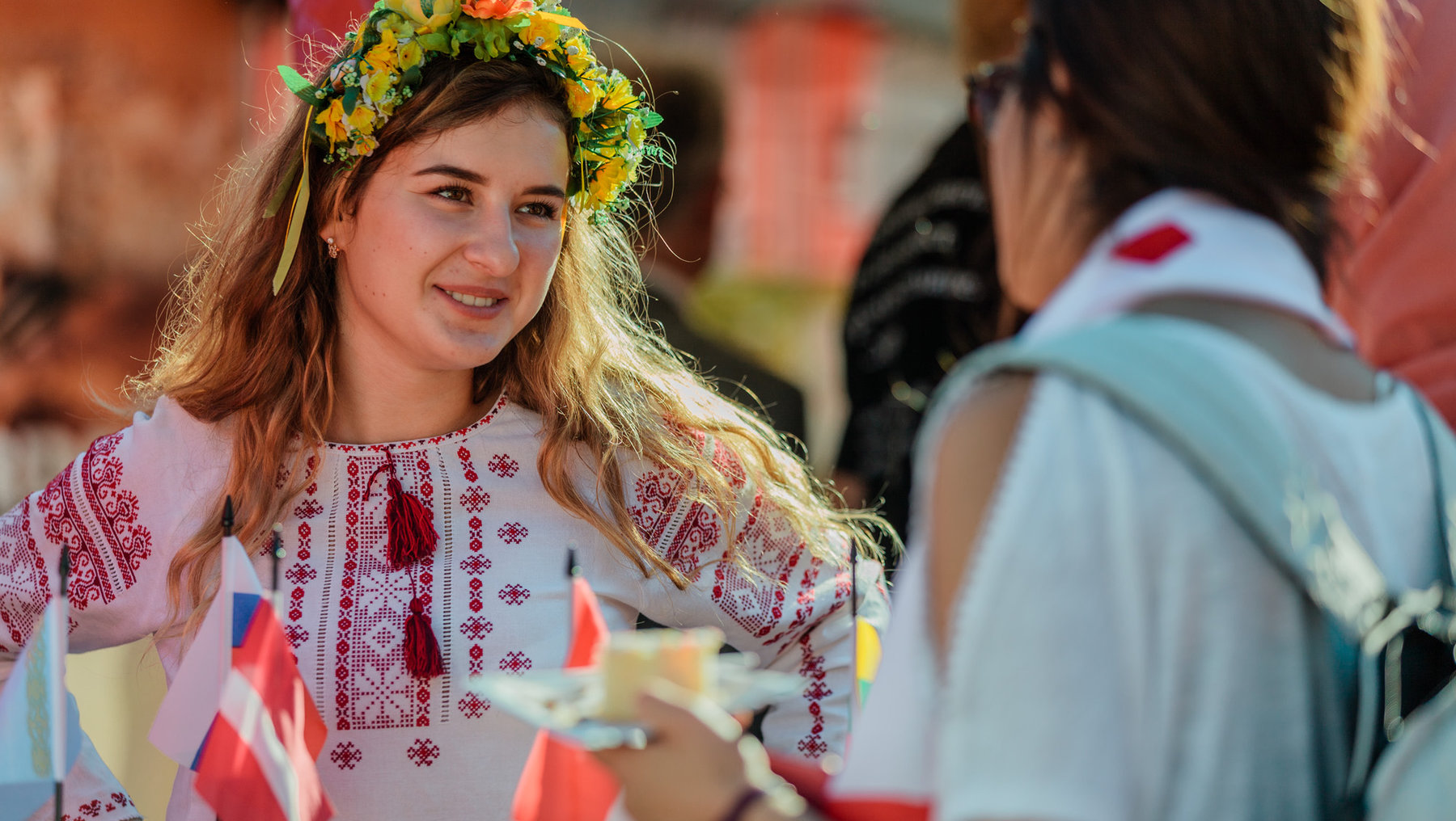 Eine lächelnde Frau mit einem Blumenkranz aus gelben, grünen und roten Blüten auf dem Kopf, die traditionelle Stickmuster auf ihrem weißen Oberteil trägt und kleine Flaggen hält.