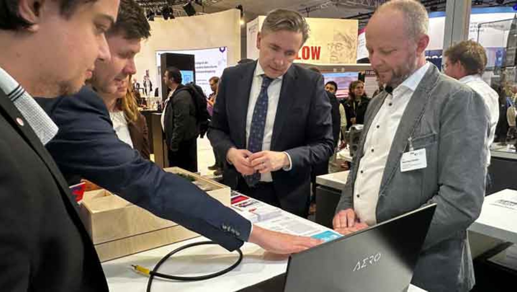 Drei Männer auf einer Messe: Einer hält ein Dokument, ein weiterer zeigt auf einen Laptop mit der Aufschrift „Aero 14“, während im Hintergrund weitere Besucher und Infostände zu sehen sind.