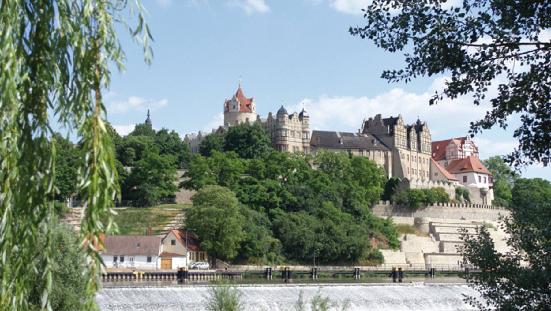 Ein malerisches Schloss auf einem Hügel, umgeben von üppigem Grün. Im Vordergrund fließt ein Fluss, und einige Gebäude sind am Ufer zu sehen. Der Himmel ist teilweise bewölkt, und Bäume rahmen das Bild ein. Die Szene vermittelt eine friedliche, historische Atmosphäre.