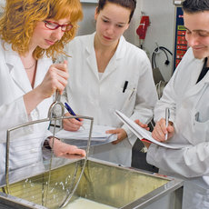 Drei Wissenschaftler in weißen Laborkitteln führen ein Experiment durch. Eine Person hält eine Pipette über einen Behälter mit Flüssigkeit, während die anderen beiden Notizen machen und die Prozedur beobachten. Sie arbeiten in einem Labor mit verschiedenen Geräten im Hintergrund.