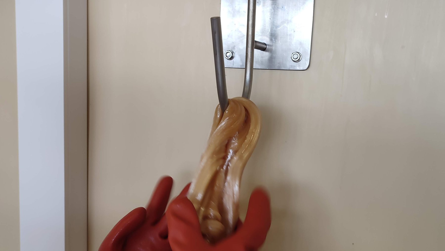 Eine Hand in einem roten Handschuh greift nach einem glänzenden, gebratenen Hotdog, der an einem Metallhaken an der Wand hängt. Der Hotdog ist in Klarsichtfolie verpackt.
