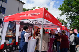 Infostand der Hochschule Anhalt mit rotem Zelt, an dem mehrere Personen stehen und Informationsmaterial zu Studiengängen wie „Game Design“ und „Game Development“ entnehmen. Website-Adresse: www.hs-anhalt.de.