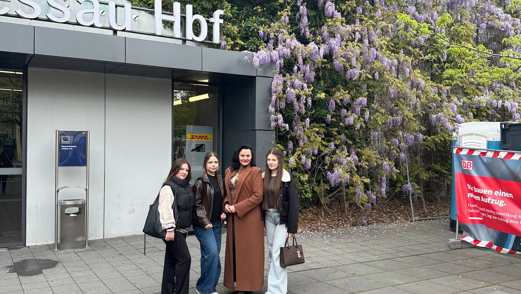 Vier Personen stehen lächelnd vor einem Bahnhofsgebäude mit dem Namen „Hauptbahnhof“, umgeben von einer blühenden, üppigen Wisteria-Pflanze in lila Blüten.