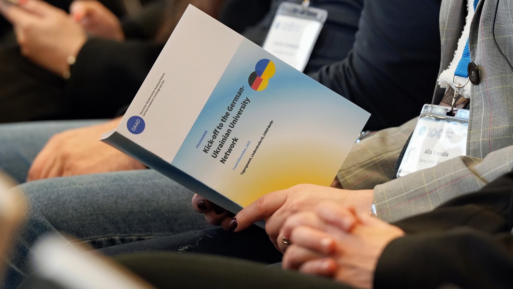 Eine Person hält ein Informationsblatt mit dem Titel "Network to the European University Network". Das Blatt zeigt ein farbenfrohes Design mit einem blauen und gelben Hintergrund. Die Person trägt einen Namensschild und sitzt in einem Raum mit anderen Menschen.