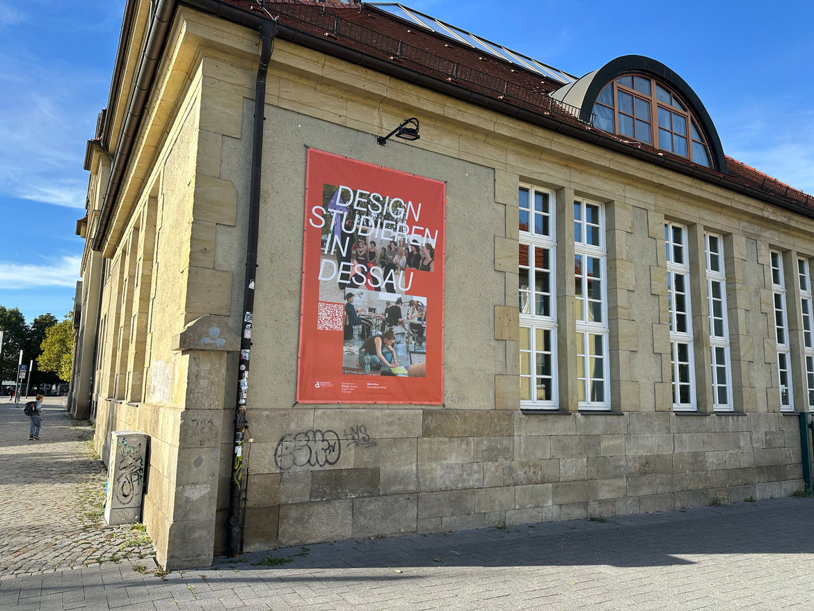 Plakat an historischem Gebäude mit der Aufschrift Design studieren in Dessau. Darunter Logo der Hochschule Anhalt. Im Hintergrund große Fenster und klassische Architektur.