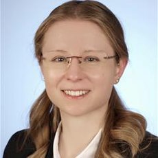 Lächelnde Person mit hellen Haaren, die zu einem Dutt gebunden sind, trägt eine Brille und ein dunkles Sakko über einem hellen Oberteil. Professionelles Porträtfoto.