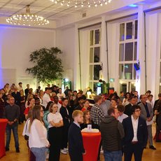 "Ein großer Raum mit vielen Menschen, die sich unterhalten. Die Gäste stehen in Gruppen zusammen und halten teilweise Getränke in den Händen. Der Raum ist hell beleuchtet mit großen Fenstern und einigen Pflanzen. Es scheint eine gesellige Veranstaltung zu sein."