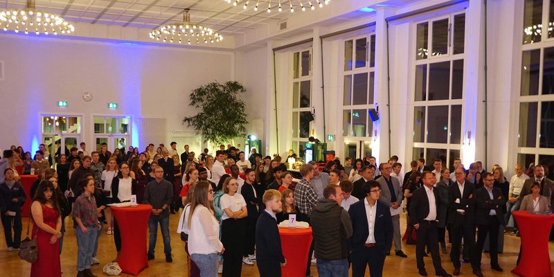 "Ein großer Raum mit vielen Menschen, die sich unterhalten. Die Gäste stehen in Gruppen zusammen und halten teilweise Getränke in den Händen. Der Raum ist hell beleuchtet mit großen Fenstern und einigen Pflanzen. Es scheint eine gesellige Veranstaltung zu sein."