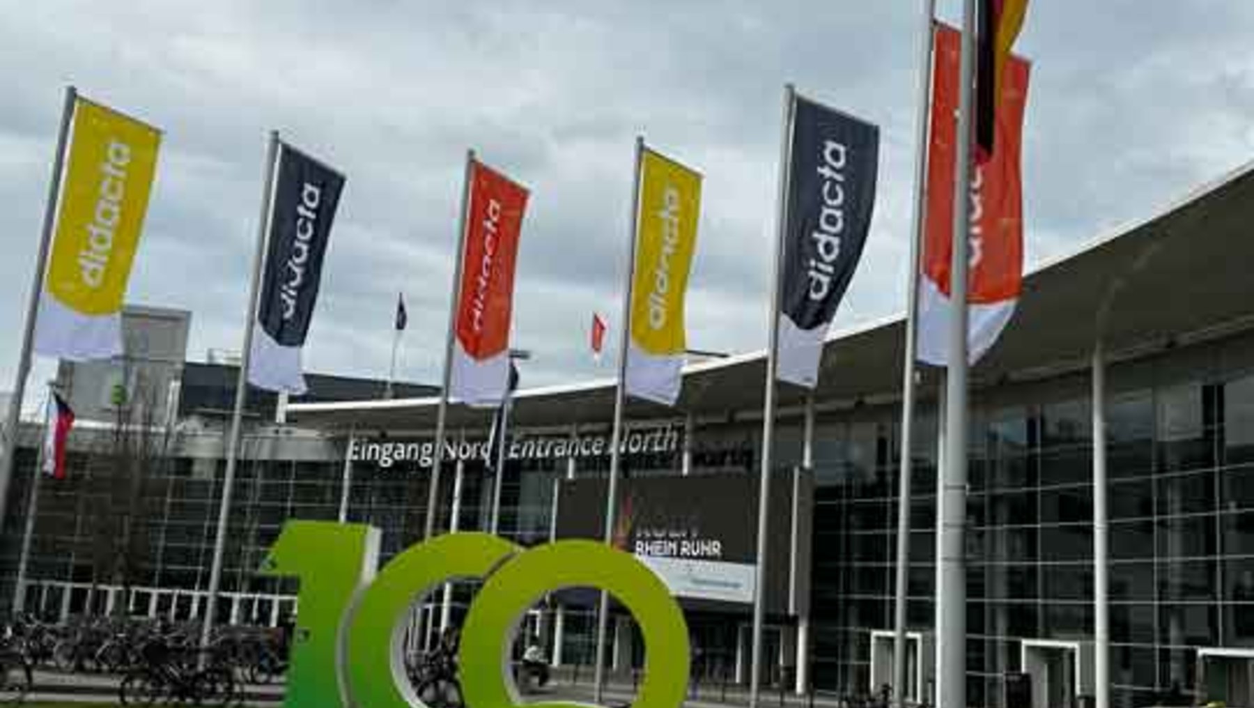 Jubiläumsfeier mit großen grünen Ziffern „100 YEARS“ vor dem Messegelände Köln, umgeben von  bunten Flaggen der Messe  „didacta“.