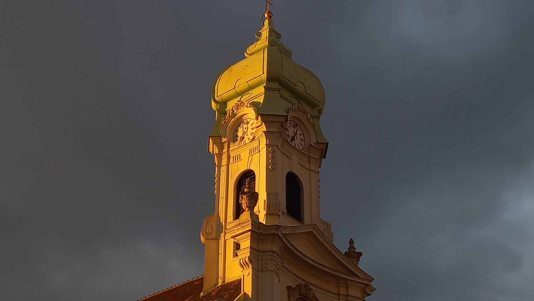 Goldgelb beleuchteter Kirchturm mit rotem Dach, zwei Glockenöffnungen und einer Uhr im oberen Bereich, vor dunklem, bewölktem Himmel.