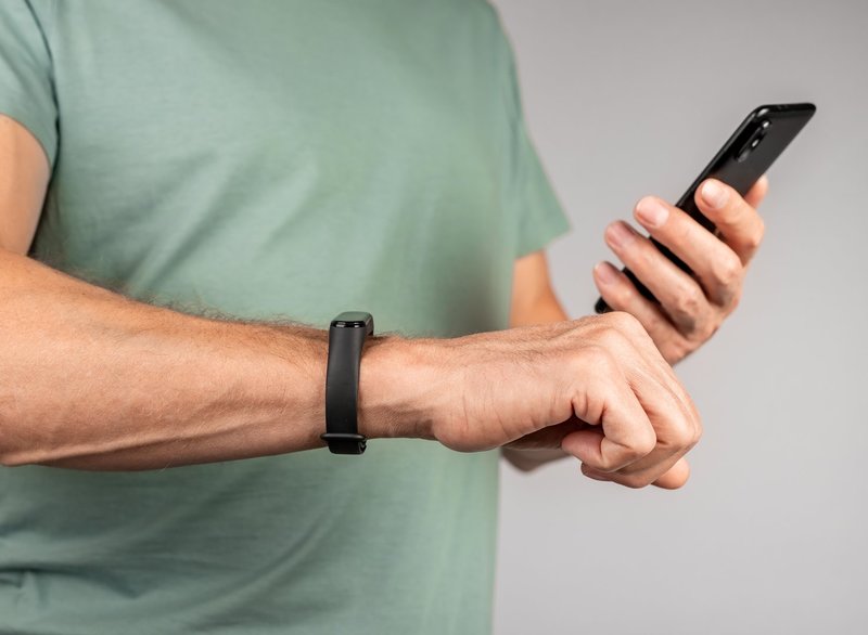 Person hält ein Smartphone in der rechten Hand und betrachtet es, während am linken Handgelenk ein schmales Fitnessarmband getragen wird.