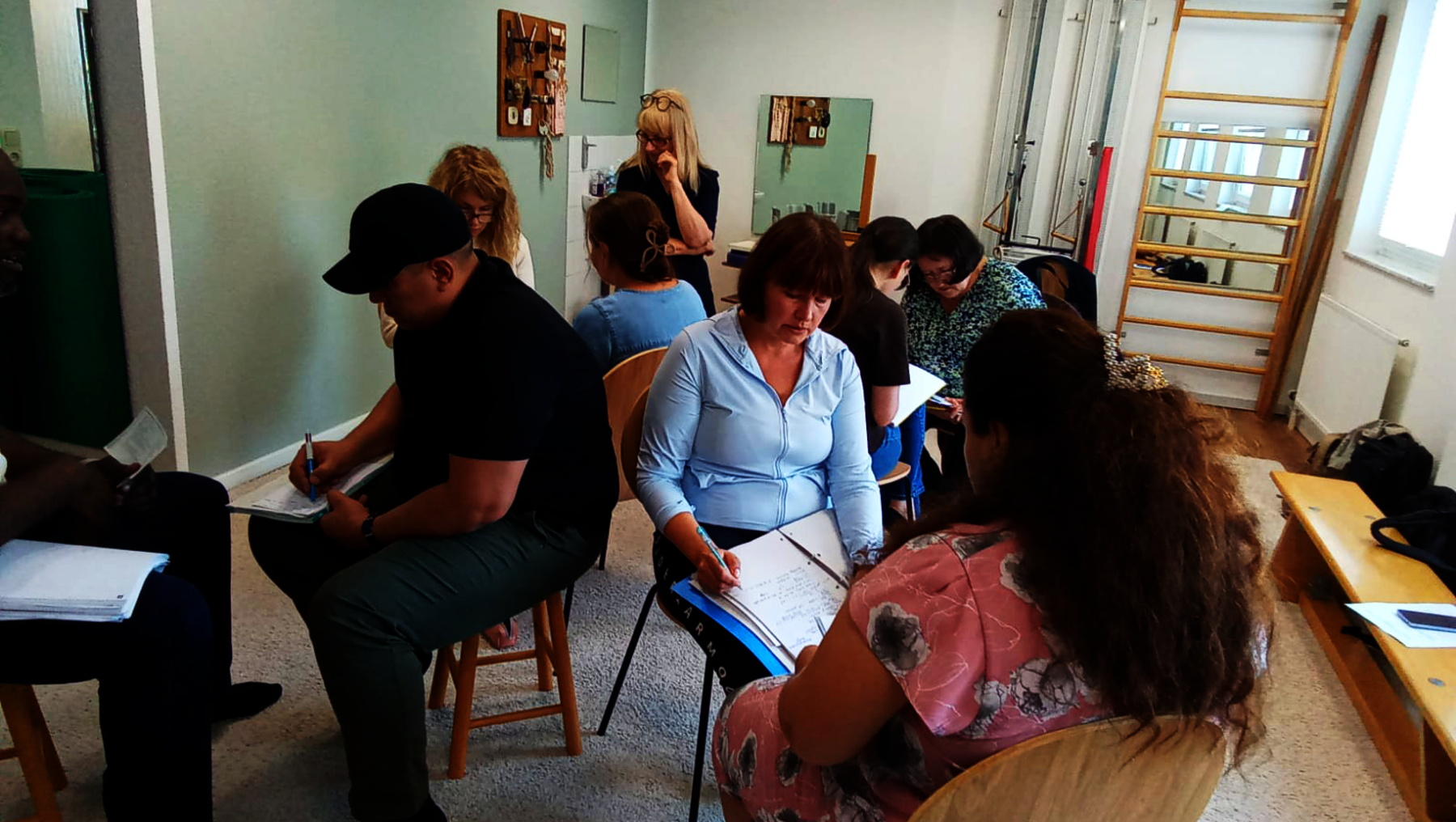 Gruppe von sieben Personen sitzt in einem hellen Raum an Tischen und scheint sich in einer Besprechung oder einem Workshop zu befinden. Eine Person hält ein Blatt Papier mit Notizen, andere hören aufmerksam zu oder schreiben mit.