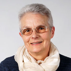 A woman with short gray hair, wearing glasses, a beige scarf, and a dark top, smiling at the camera.